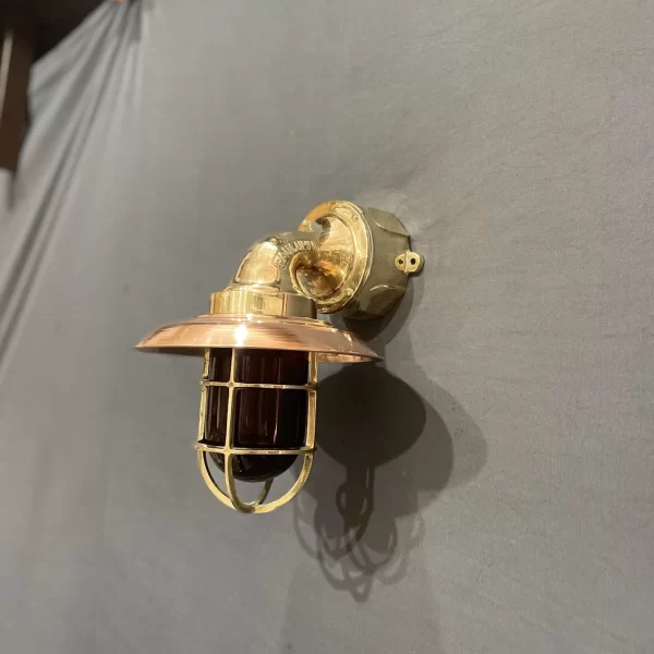 Brass and copper nautical wall sconce with green glass cage light. Vintage ship bulkhead light with green lens and brass cage. Handmade maritime wall lamp in brass and copper with green glass shade. Industrial coastal wall sconce with cage-protected green glass bulb cover.
